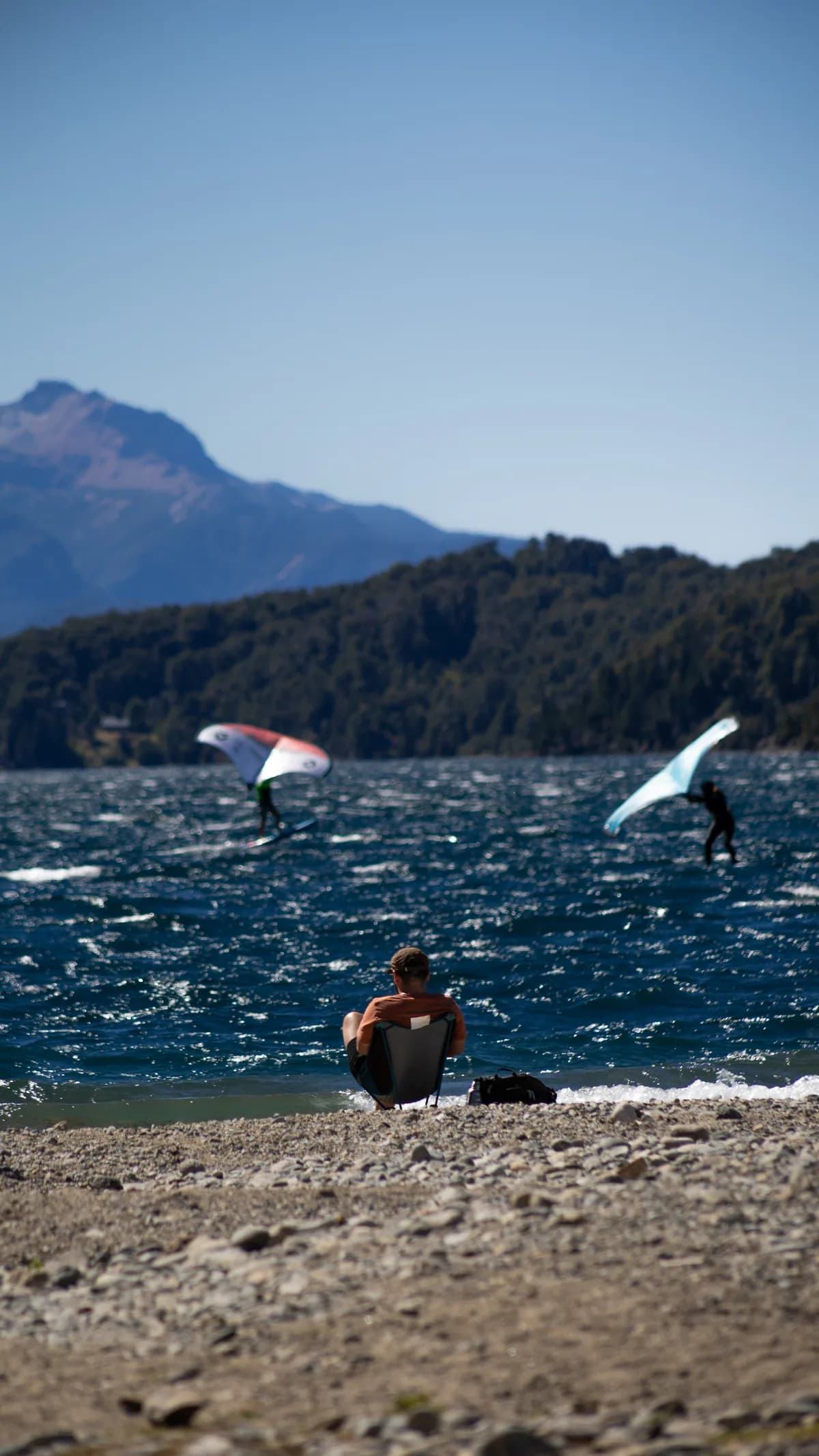 Man sunbathing on a lake shore with three people doing wingfoil.