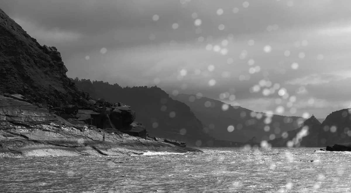 Water drops from waves hitting a boat, with a fisherman on the shore and mountains in the background