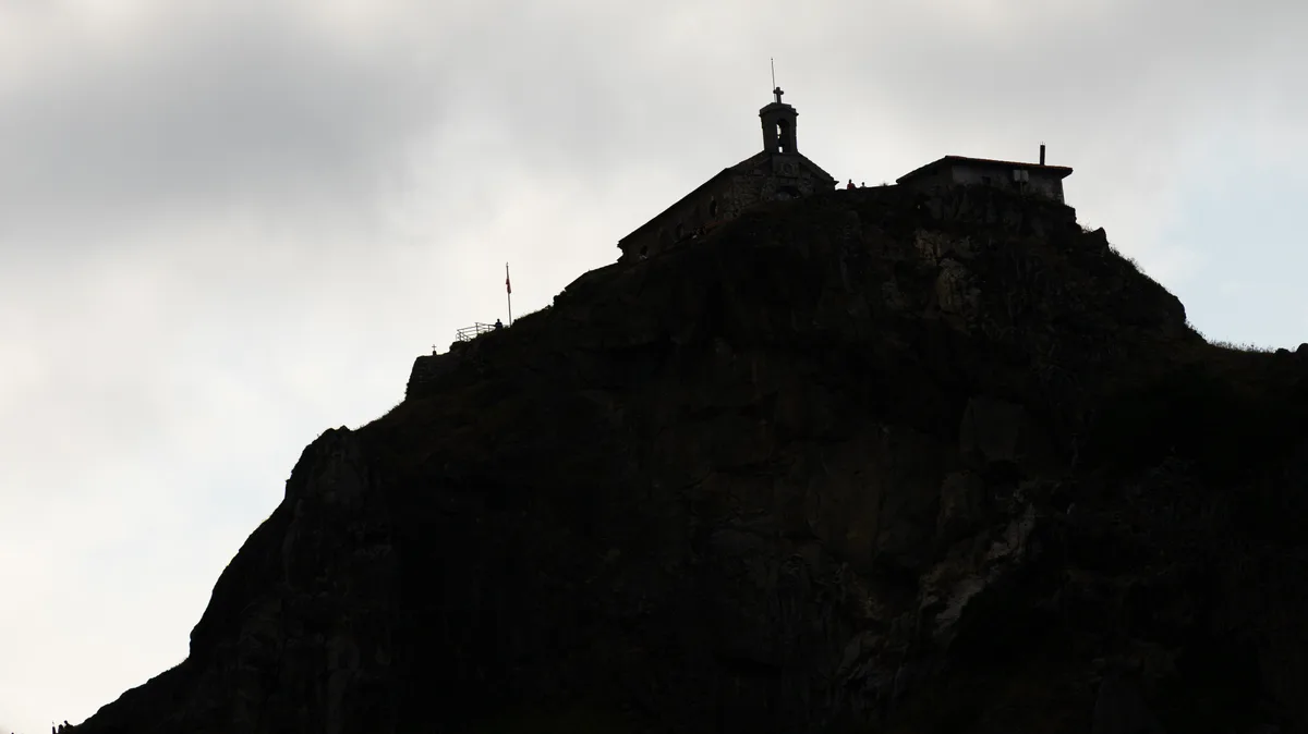 A stone chapel with a bell tower is silhouetted against bright sky atop a rocky summit