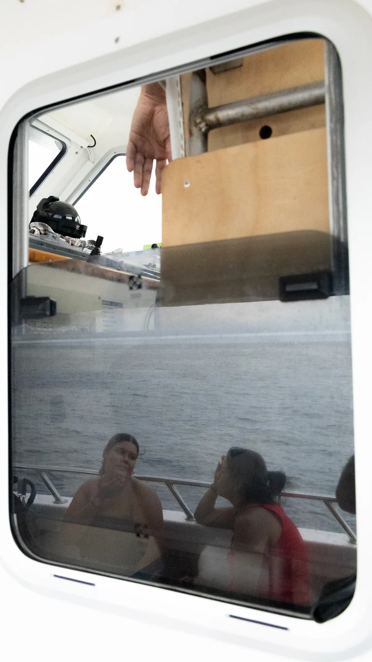 A boat window reflects gear on deck while two seated women are visible faintly through the glass