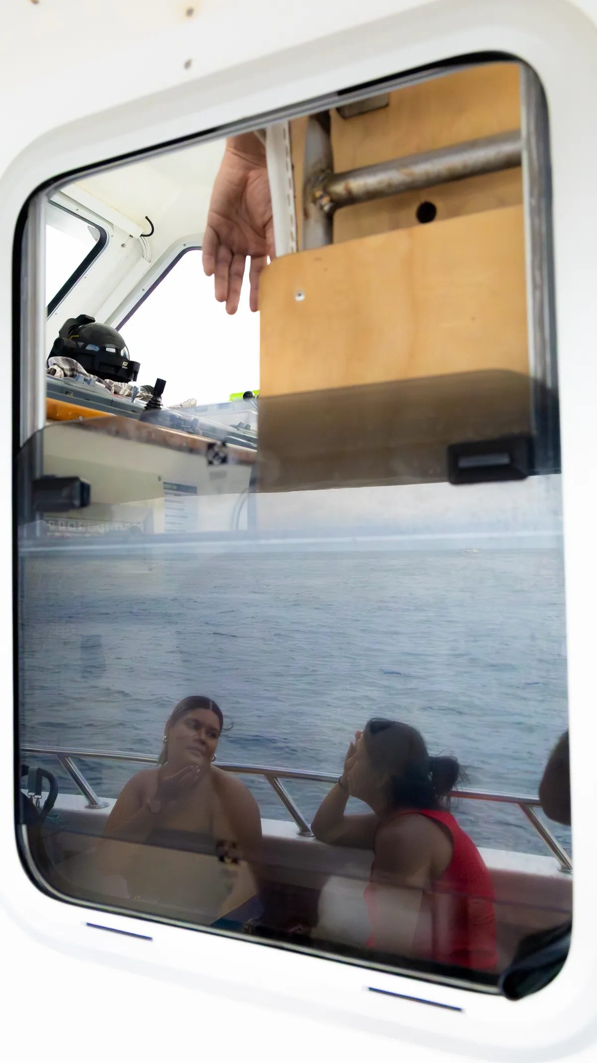 A reflection of two women chatting in the bottom half of a boat window, the top one shoting the helmsman