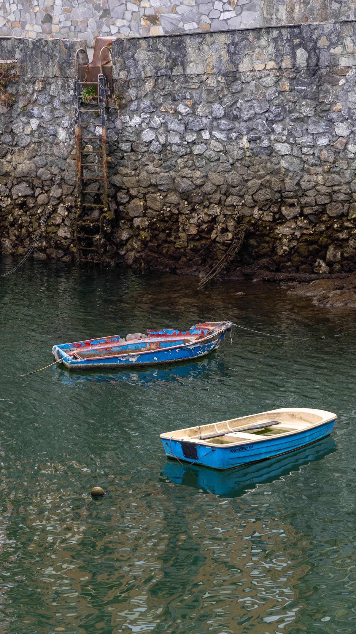 Two small weathered rowboats float on still green water beside a stone quay wall