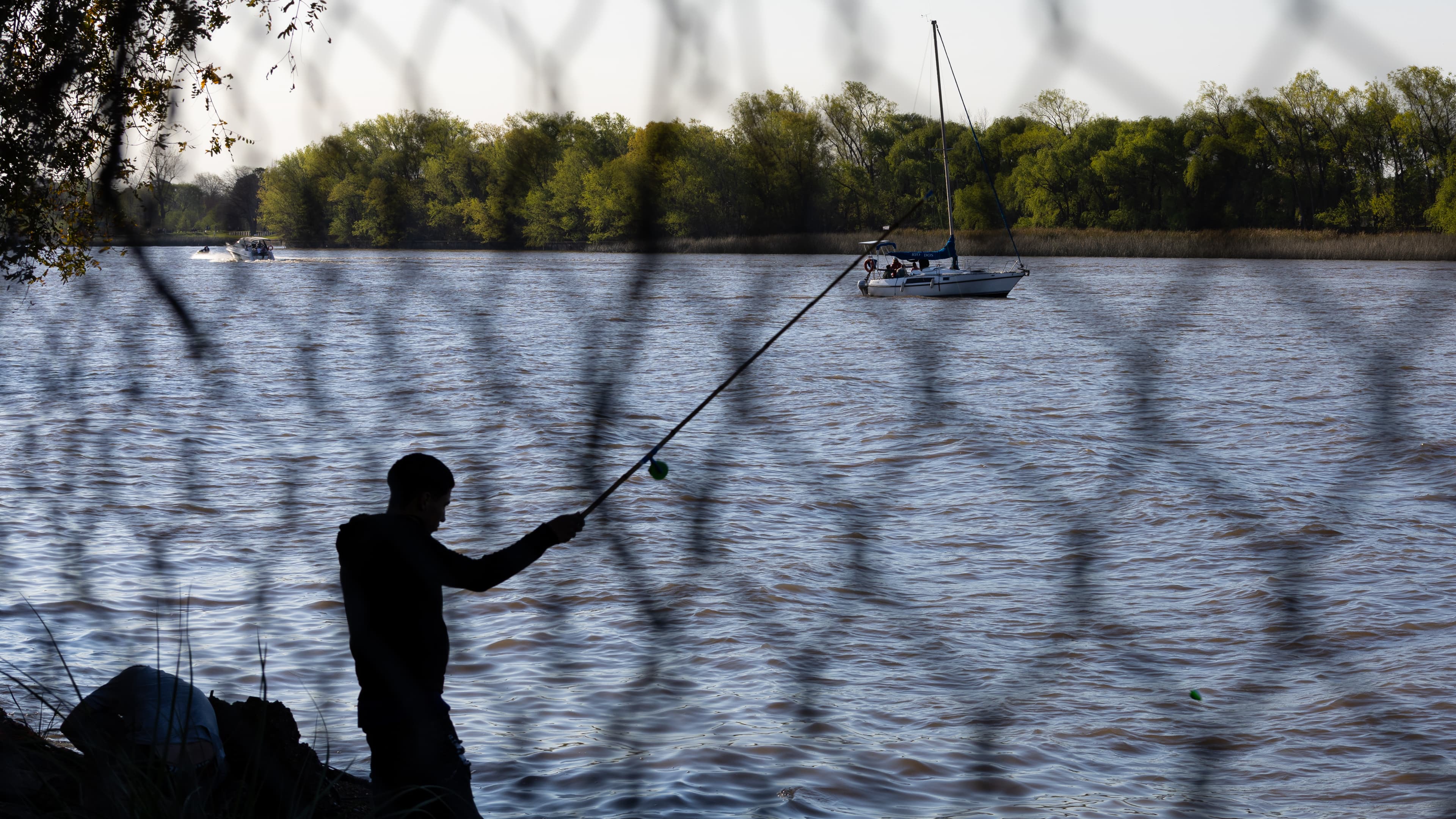 Man fishing behind a fence with sailboat passing by