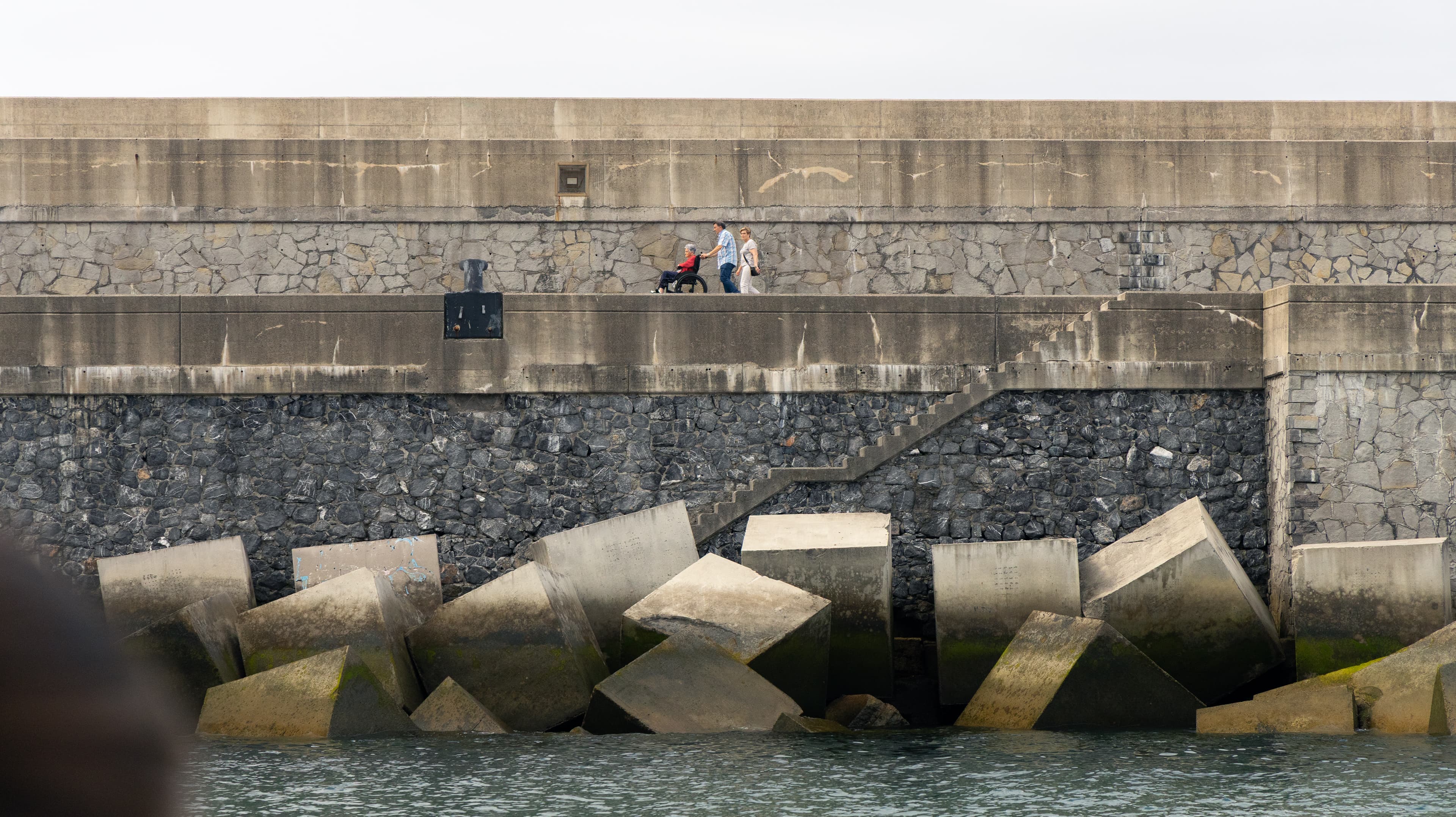 People walk along a stone breakwater above jumbled concrete tetrapods at the waterline