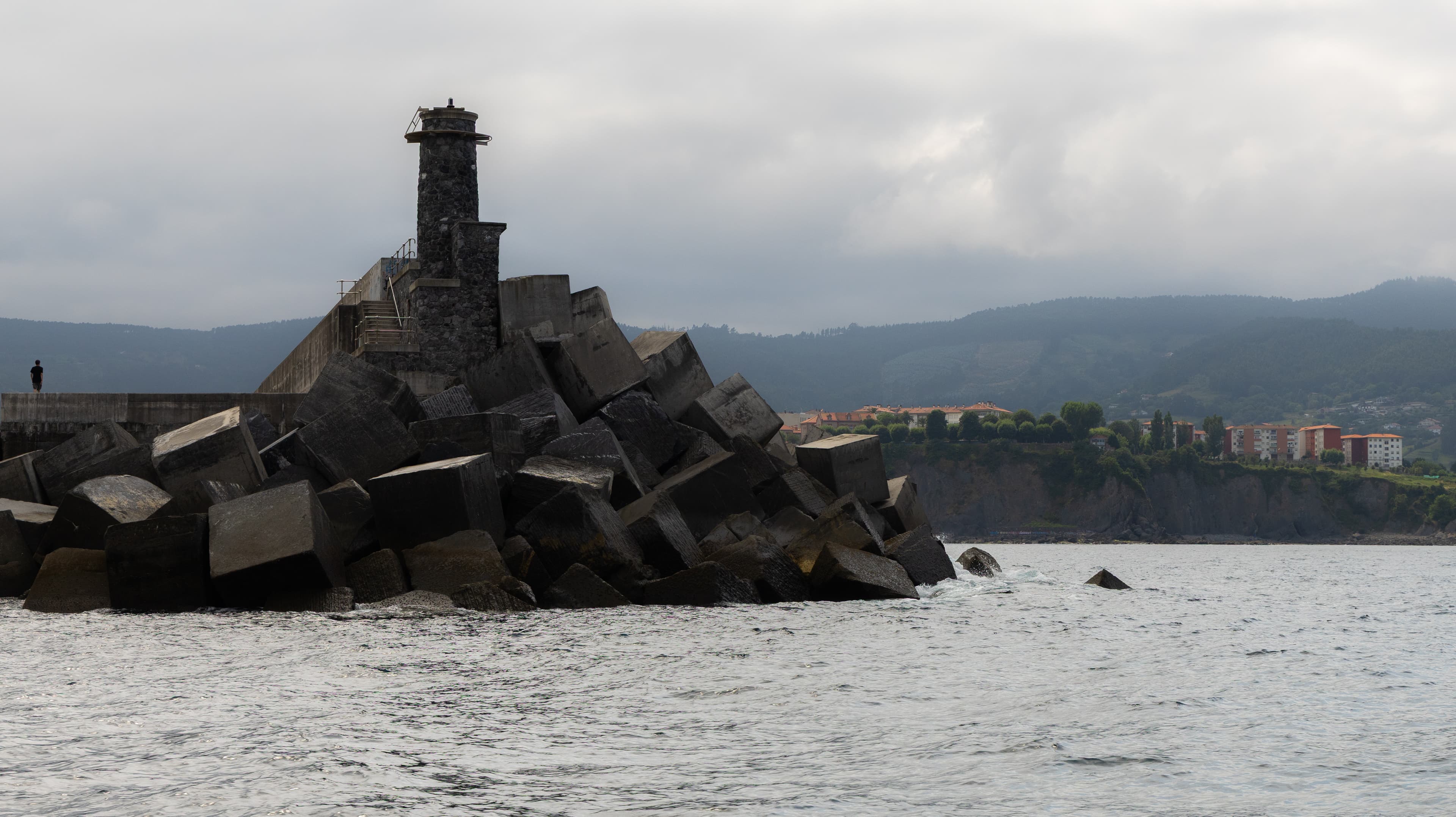 A stone lighthouse tops a jagged rock outcrop jutting into the sea near a coastal town