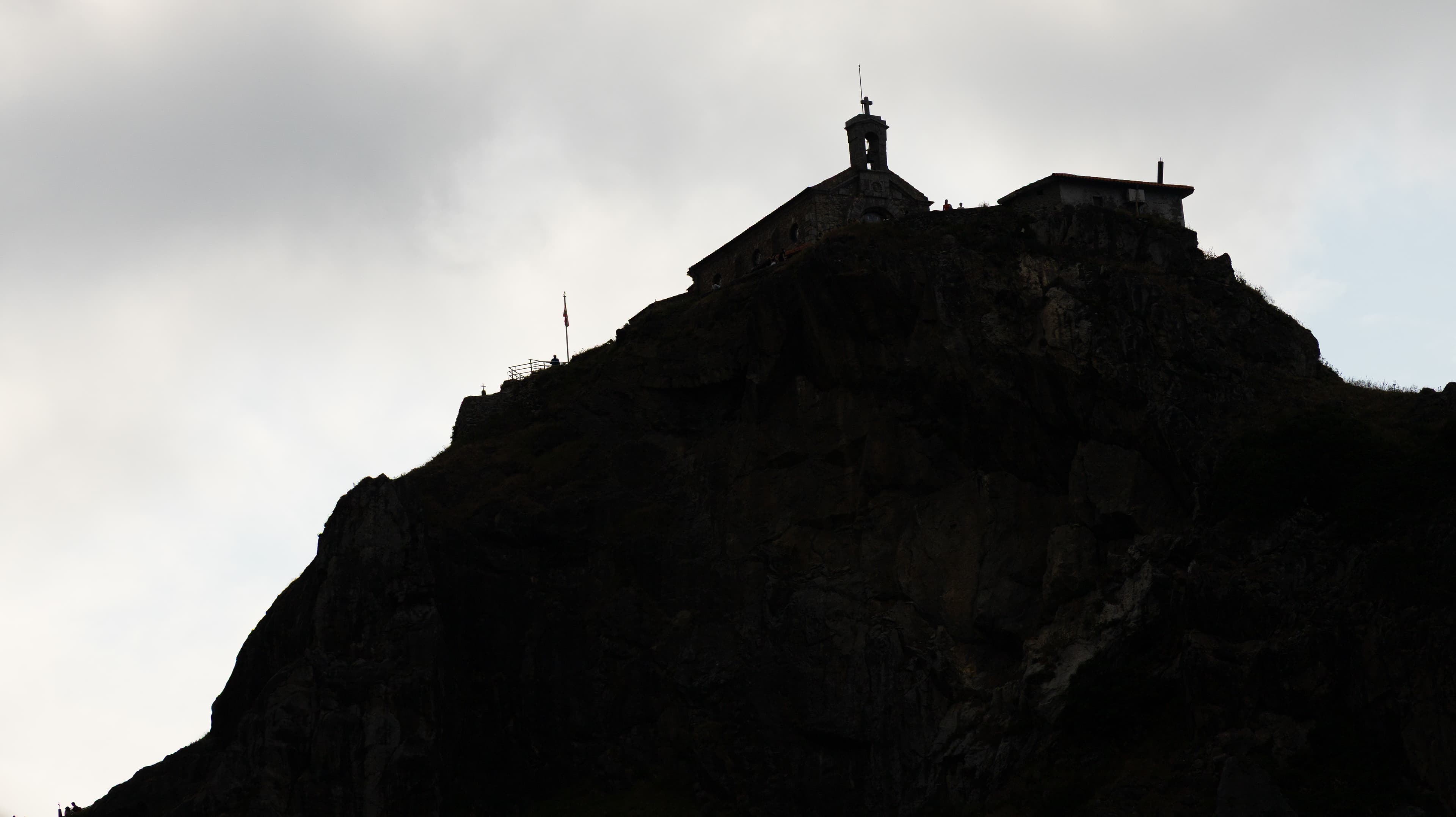 A stone chapel with a bell tower is silhouetted against bright sky atop a rocky summit