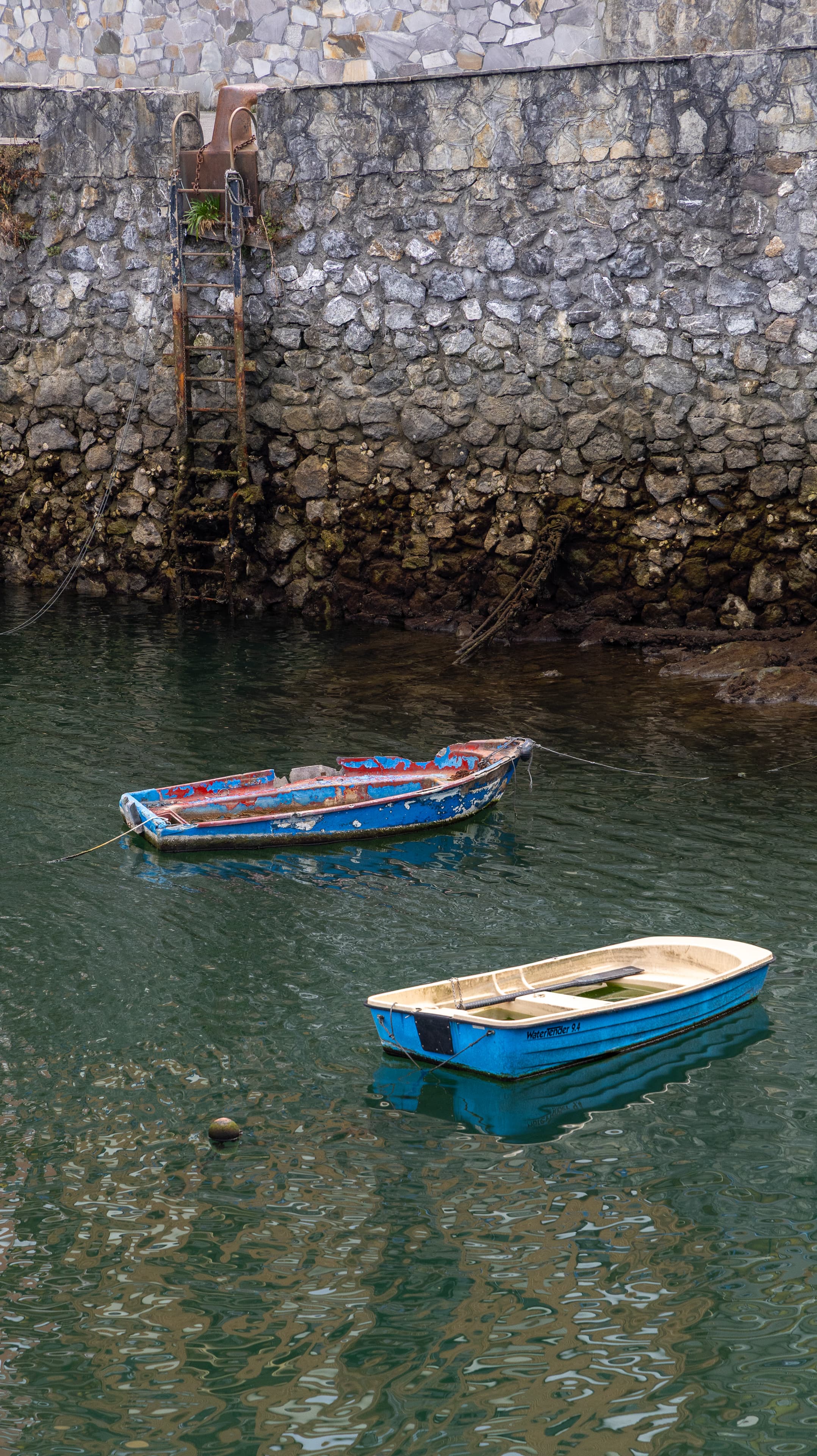 Two small weathered rowboats float on still green water beside a stone quay wall